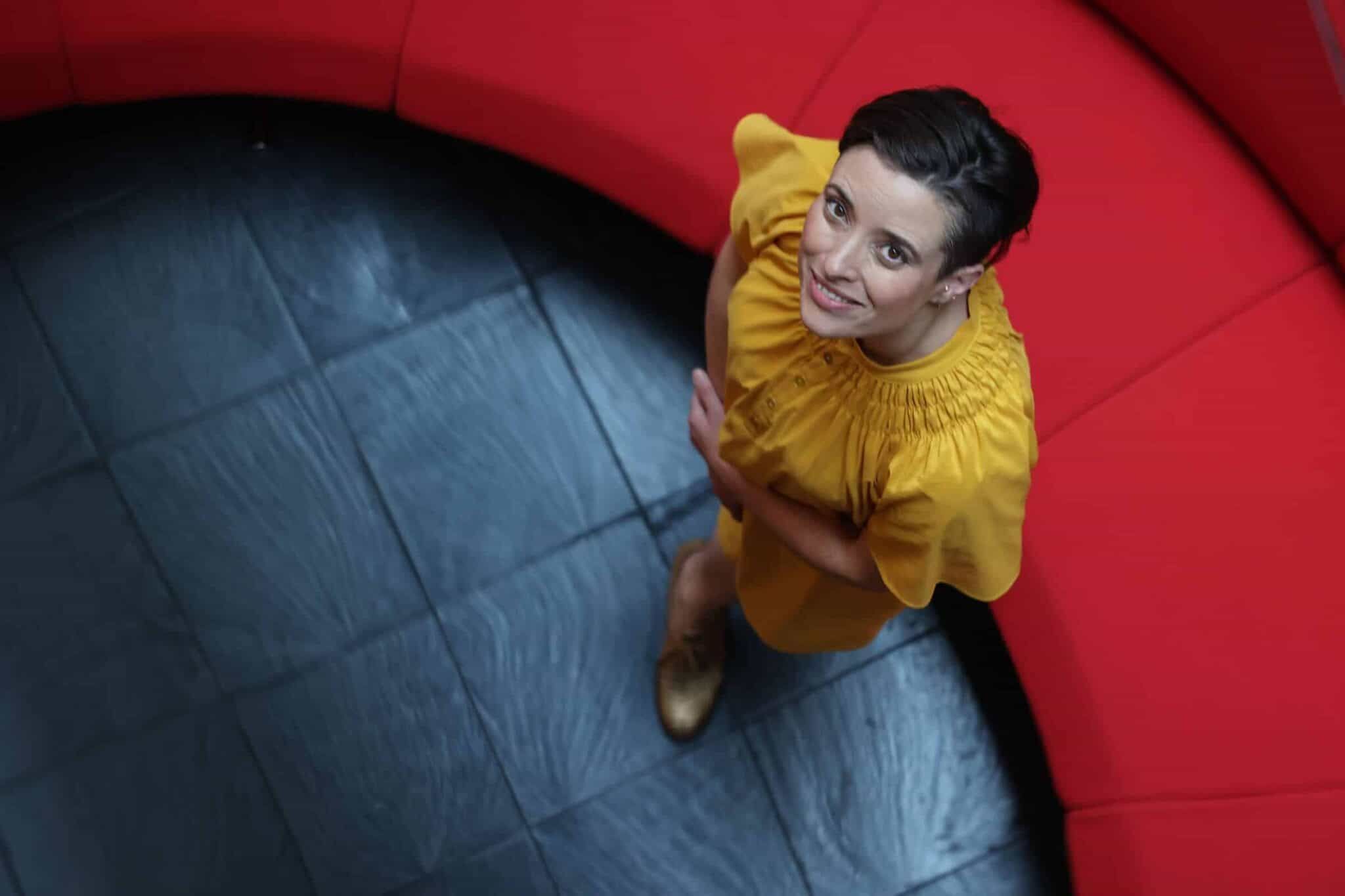 Karen Melchior in a yellow dress standing on black tiles by a red sofa looking up and smiling